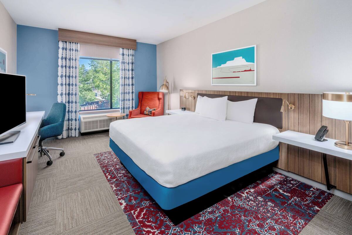 A hotel room with a large bed, a desk with a computer, a chair, and a window with curtains.