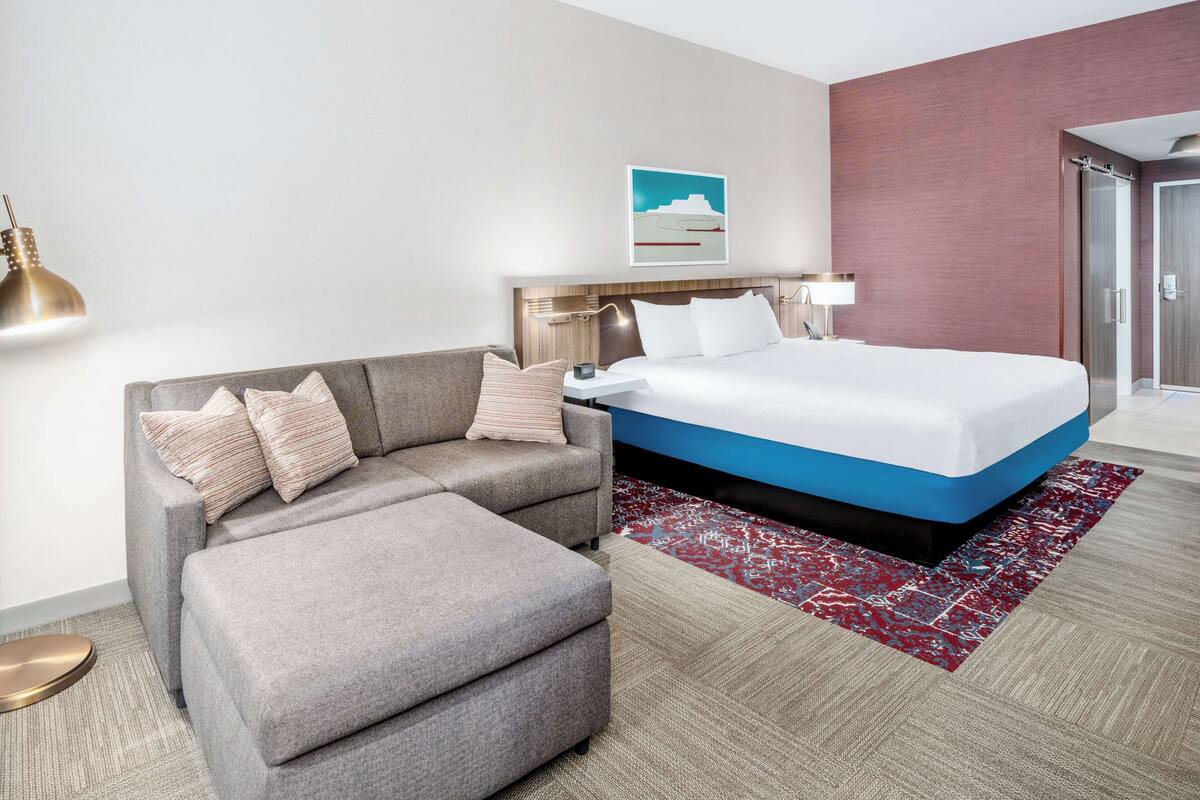 A hotel room with a large bed, a grey sofa, a small ottoman, a red patterned rug, a wall-mounted lamp, and a framed picture on the wall.