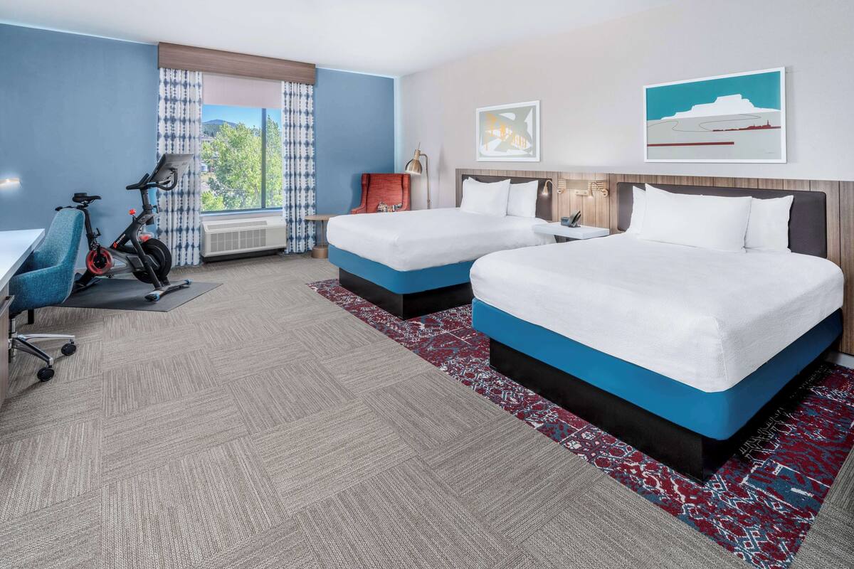 A hotel room with two beds, a desk with an exercise bike, a large window with curtains, and artwork on the walls.