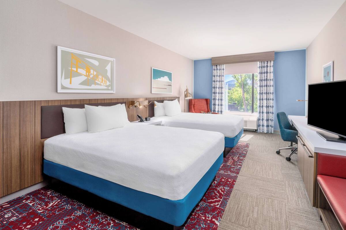 A hotel room with two beds, a desk, a TV, and a window with curtains.