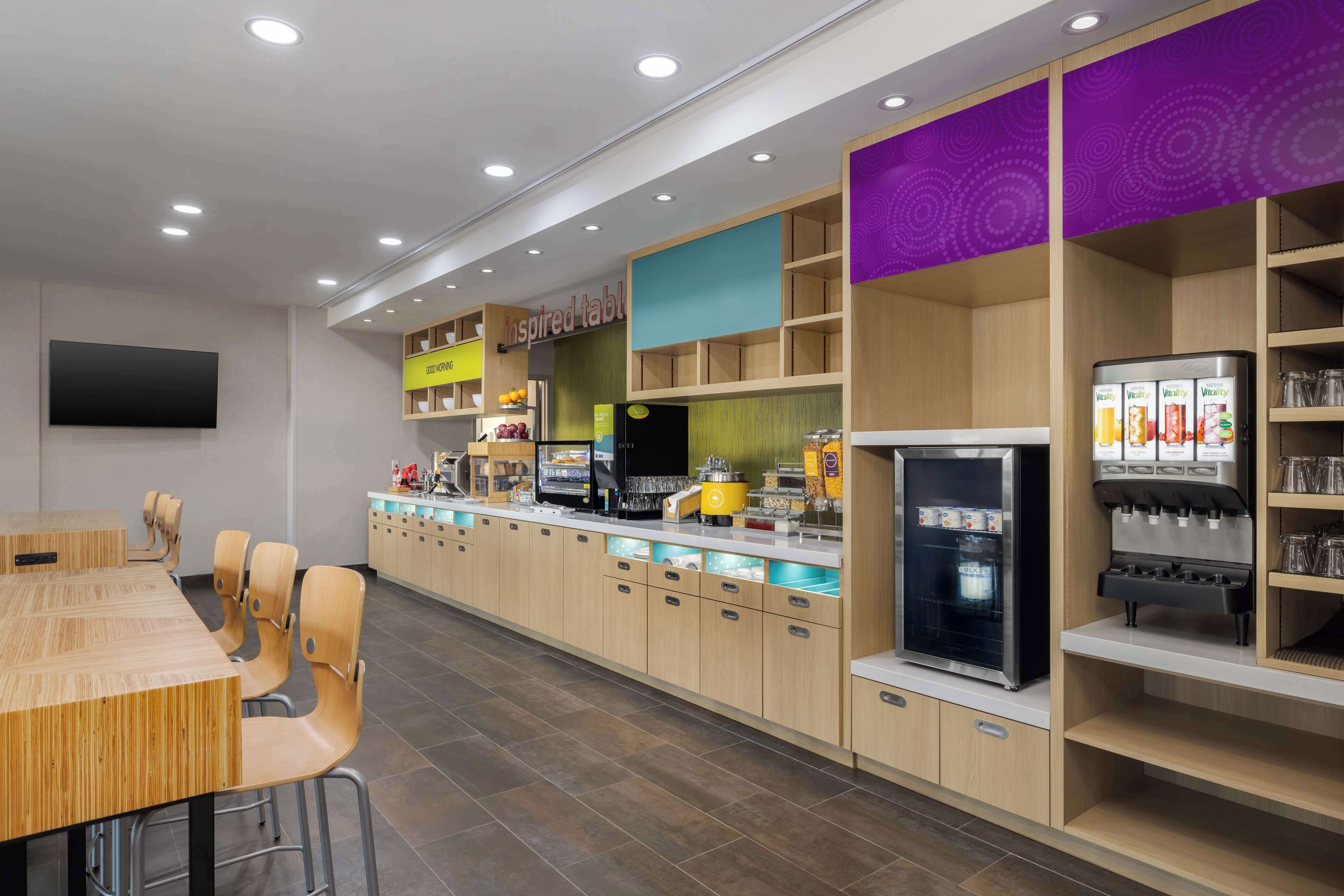 A modern restaurant with a long counter, wooden chairs, a refrigerator, and a beverage dispenser.
