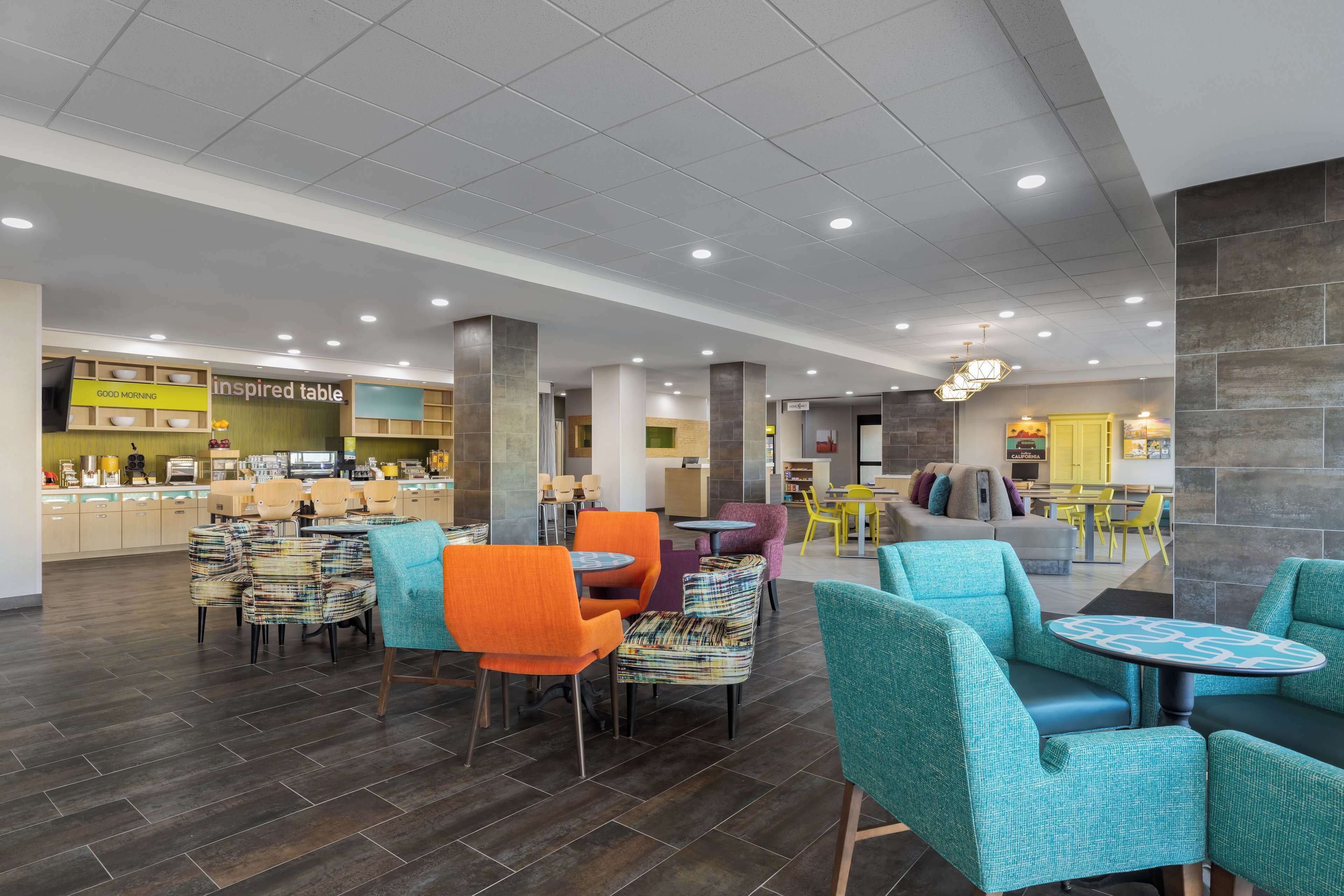 A modern hotel lobby with a variety of seating options, including colorful chairs and tables, and a visible restaurant area in the background.