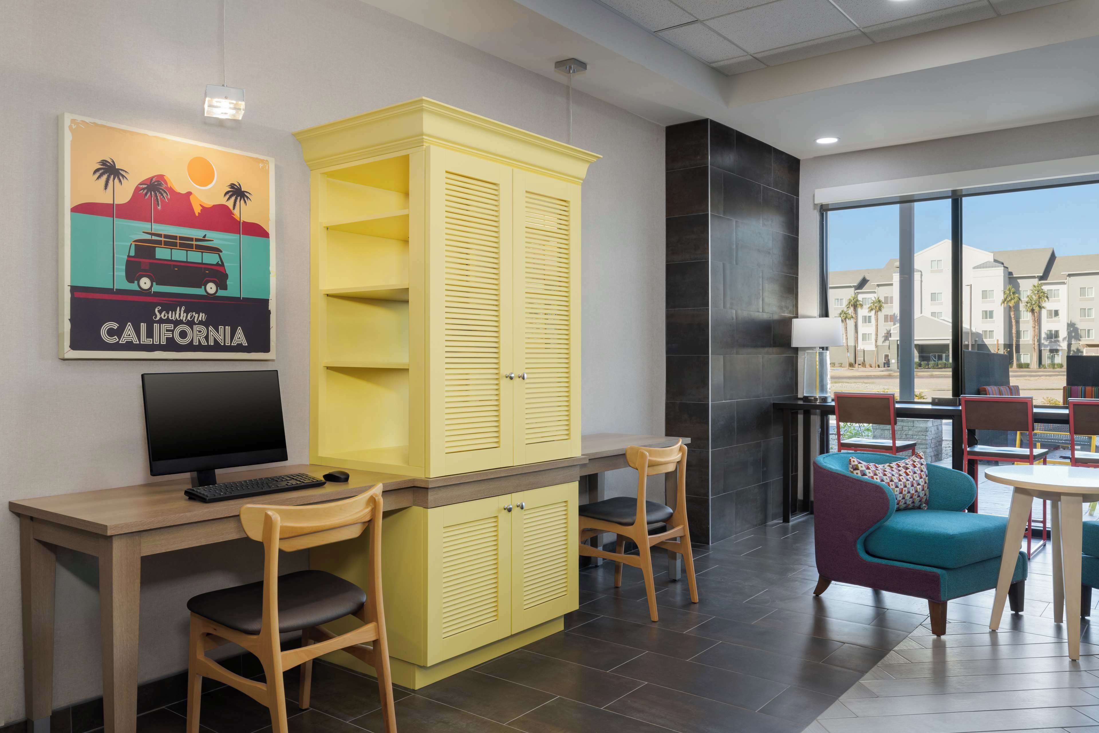 Une chambre d’hôtel moderne avec un bureau en bois, une armoire jaune, un fauteuil turquoise et une grande fenêtre donnant sur la ville.