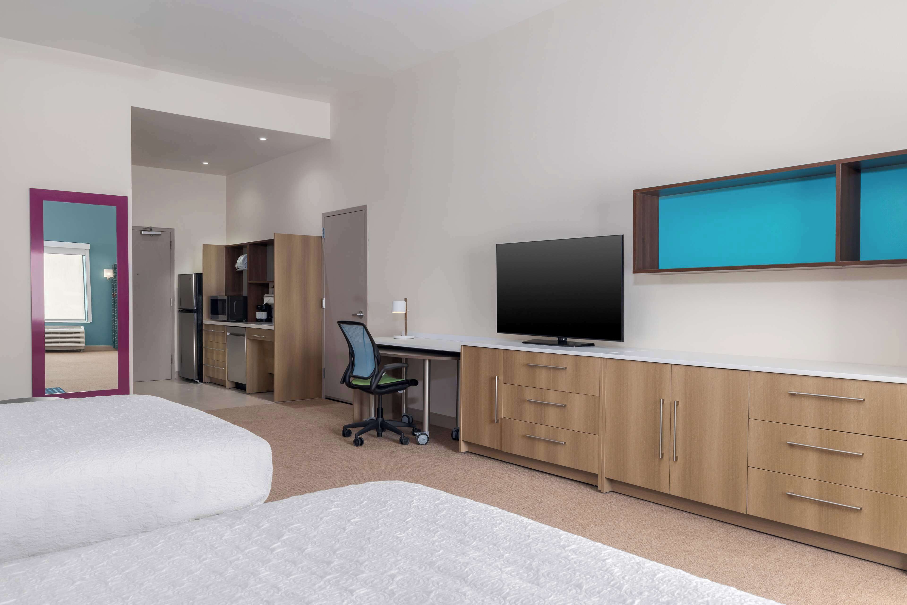 A modern hotel room with a large bed, a desk with a computer, a television, and a kitchenette.