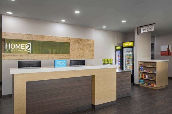En hotellobby med en receptionsdisk, en skylt med texten ”HOME 2”, en skylt med texten ”Hilton Honors”, ett ”riktigt kallt” kylskåp och en hylla med snacks.