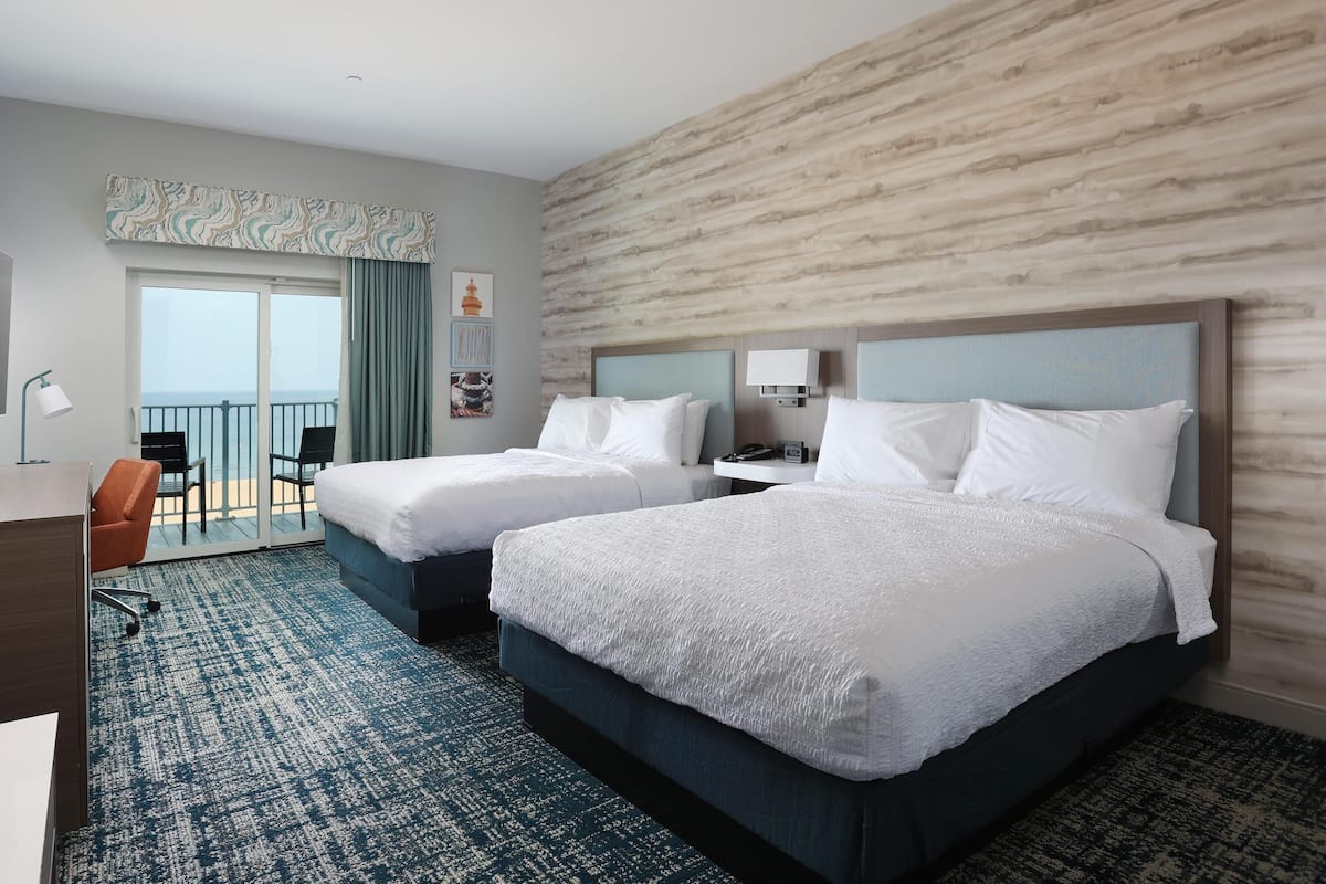 A hotel room with two beds, a desk, a chair, a balcony with a view of the sea, and a wall with a wood-like texture.
