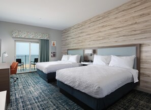 A hotel room with two beds, a desk, a chair, a balcony with a view of the sea, and a wall with a wood-like texture.