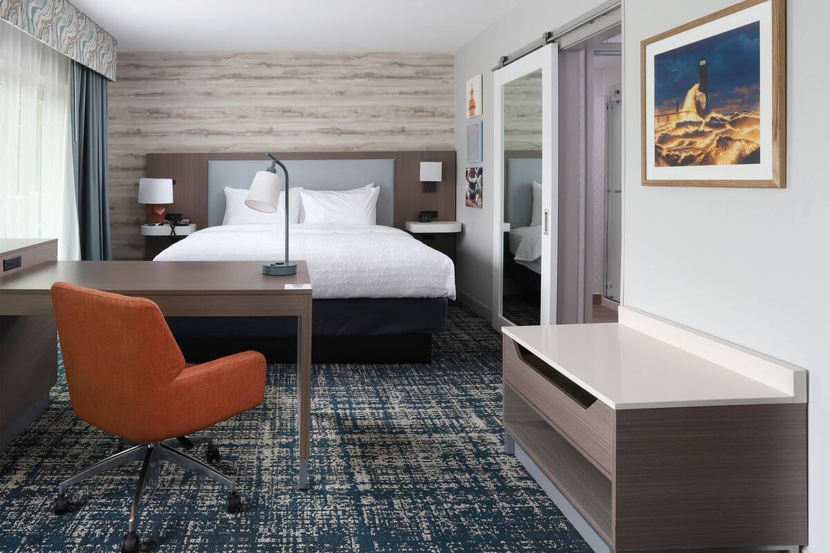 A hotel room with a bed, desk, chair, and a painting on the wall.