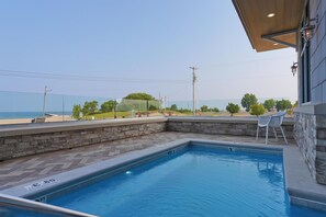A swimming pool with a stone border, a view of the beach, and a covered patio area with chairs.