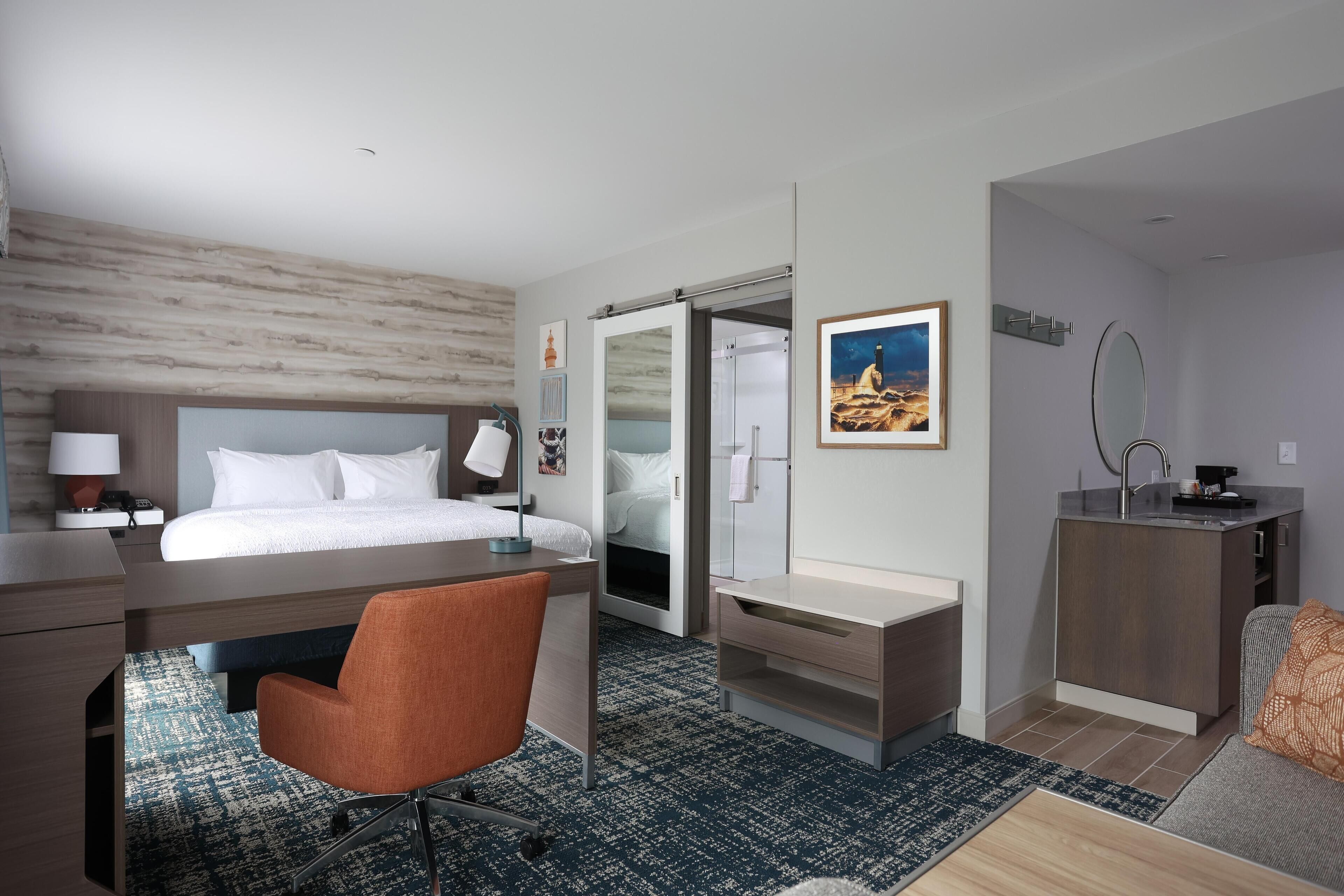 A modern hotel room with a bed, desk, chair, and a small kitchenette.