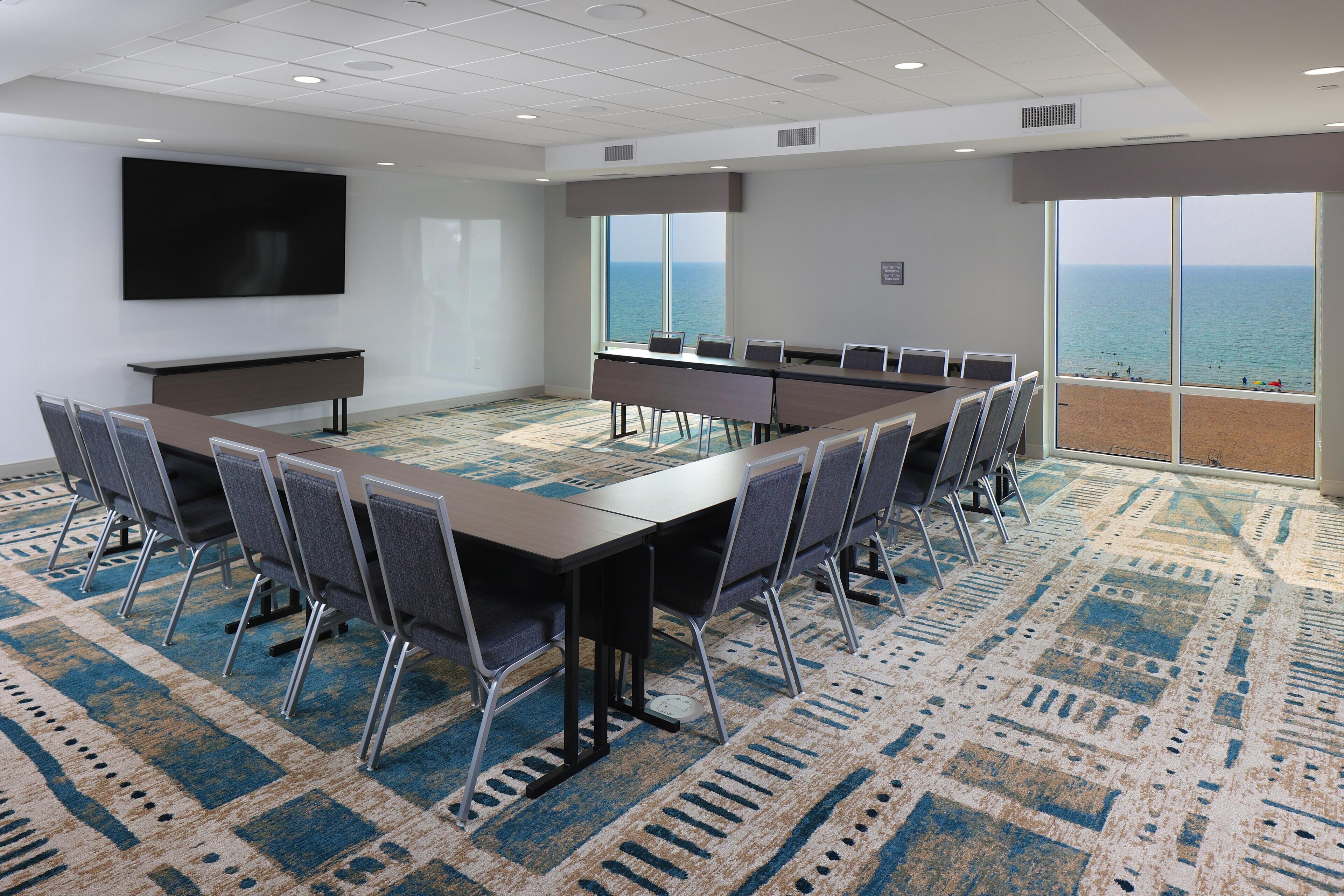 A conference room with a long table, chairs, a TV screen, and large windows with a view of the ocean.