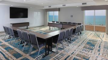 Un salón de conferencias con una larga mesa, sillas, una pantalla de televisión y grandes ventanales con vista al océano.