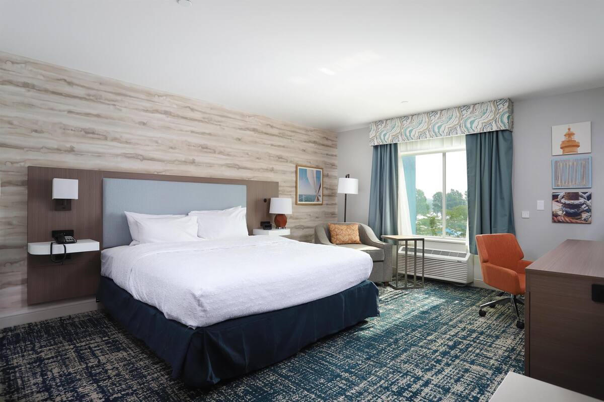 A hotel room with a large bed, a desk, a chair, a small table, and a window with curtains.