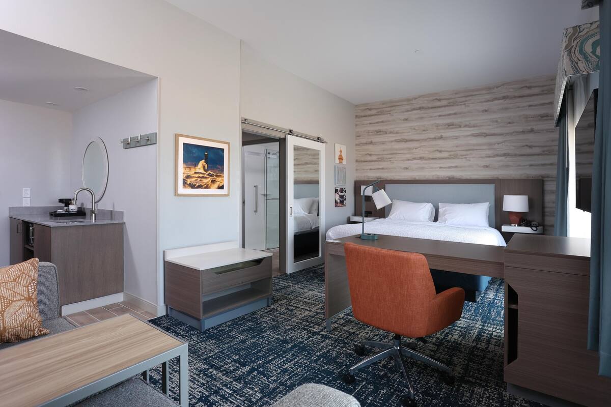 A hotel room with a bed, desk, chair, and a small kitchenette.