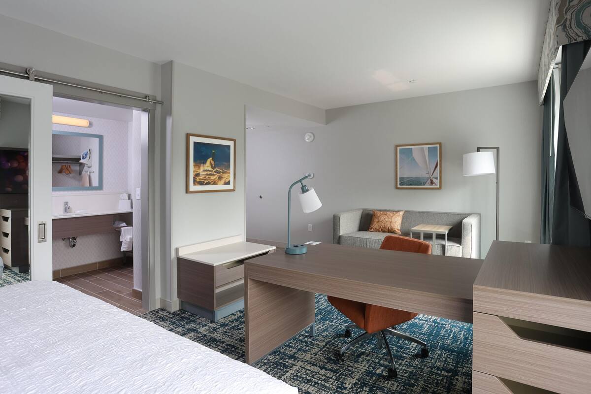 A hotel room with a bed, a desk, a chair, a sofa, a lamp, and artwork on the walls.