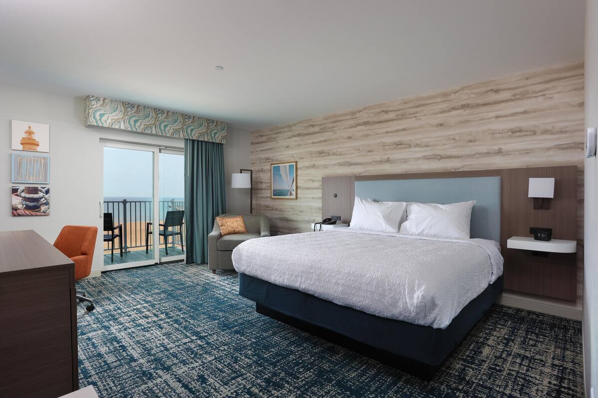 A hotel room with a large bed, a desk, a chair, a balcony with a view, and a wall with a patterned design.