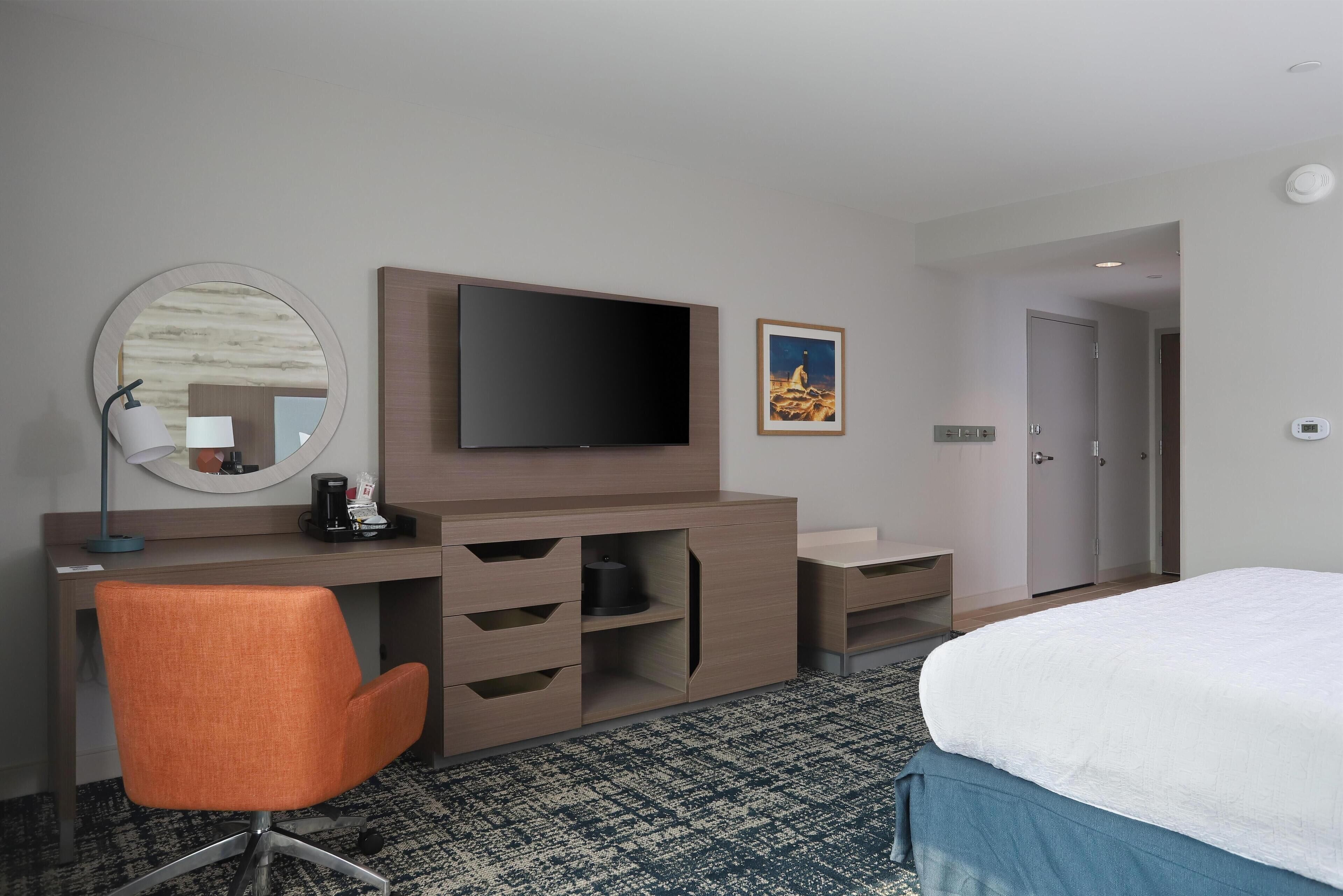 Hotelkamer met een bed, een bureau met flatscreen-tv, een stoel, een ronde spiegel, een koffiezetapparaat en een lijst aan de muur.