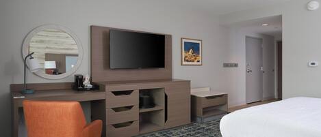 Hotelkamer met een bed, een bureau met flatscreen-tv, een stoel, een ronde spiegel, een koffiezetapparaat en een lijst aan de muur.