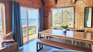 A wooden cabin with a large sliding glass door leading to a balcony, a dining area with a wooden table and chairs, and a view of the ocean. - Ocean Front A Frame with Spectacular Views! Dog Friendly! Fireplace! (Florence)