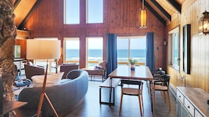 A cozy cabin with a wooden interior, large windows overlooking the ocean, a dining area with a table and chairs, and a stone wall. - Ocean Front A Frame with Spectacular Views! Dog Friendly! Fireplace! (Florence)