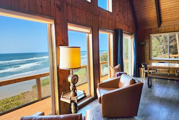 A room with large windows overlooking the ocean, a wooden interior, and a table with chairs.