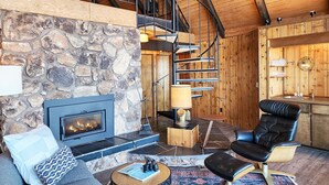 A cozy living room with a stone wall, a fireplace, a black leather chair, a round coffee table, and a spiral staircase leading to an upper level. - Ocean Front A Frame with Spectacular Views! Dog Friendly! Fireplace! (Florence)