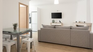 Un salon moderne avec une table à manger en verre, des tabourets blancs, un canapé gris et un téléviseur à écran plat fixé au mur.