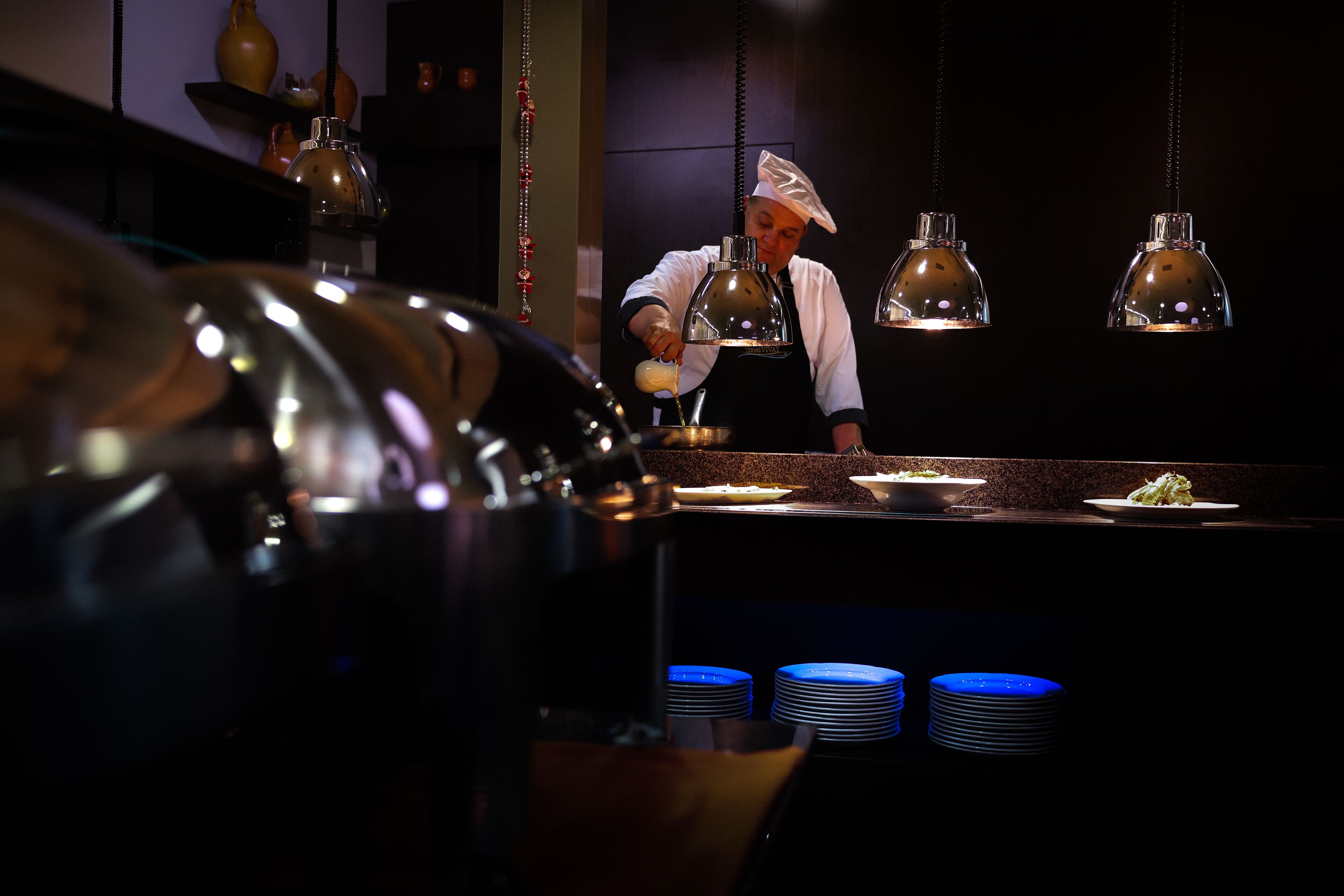 A chef preparing food in a dimly lit kitchen with hanging pendant lights and stacks of blue plates.