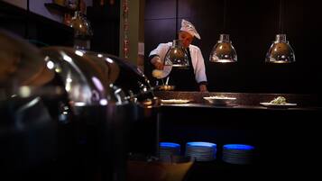 A chef preparing food in a dimly lit kitchen with hanging pendant lights and stacks of blue plates.