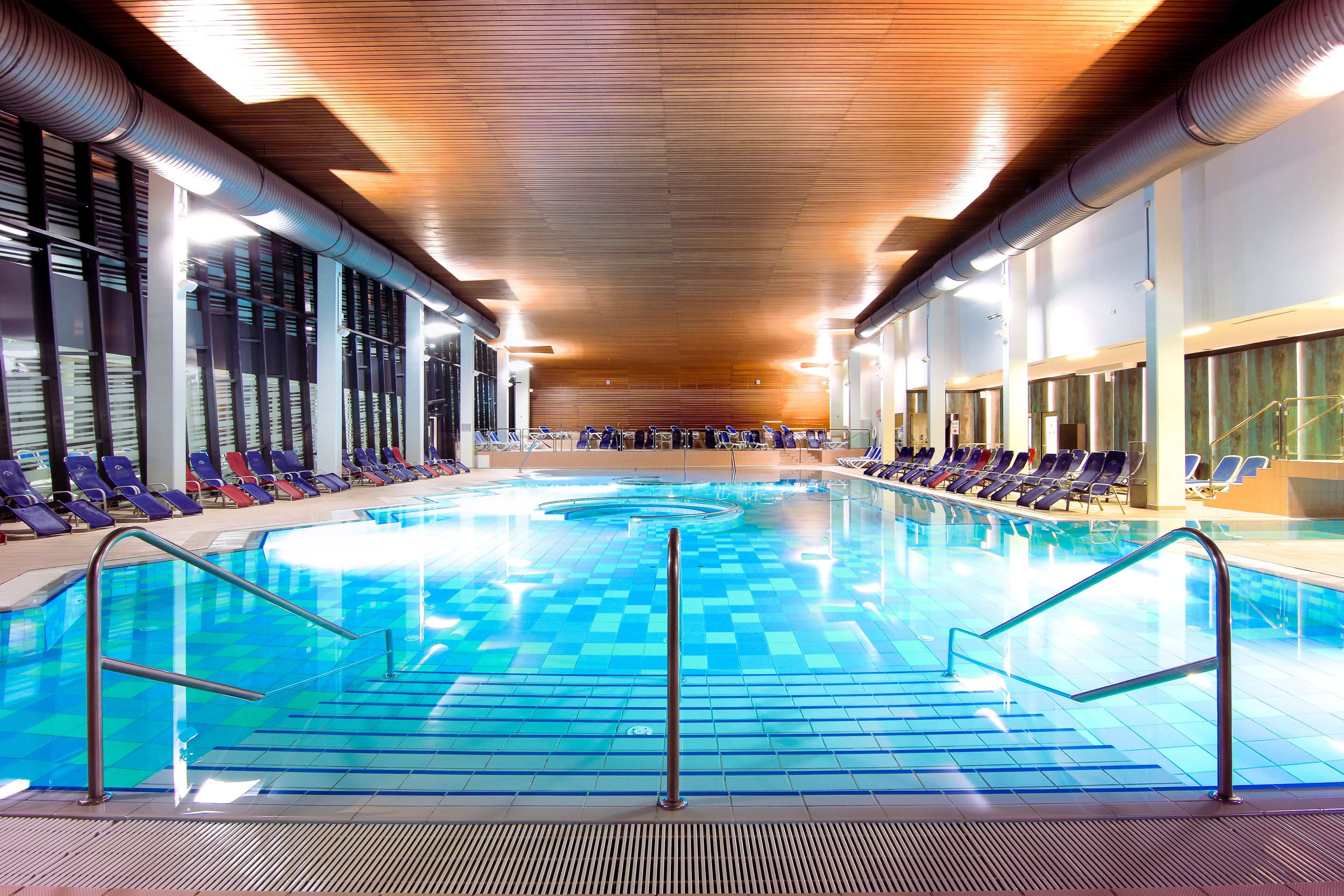 Una piscina interior con baldosas azules cristalinas, barandillas metálicas y filas de sillones de relax a lo largo de los bordes.
