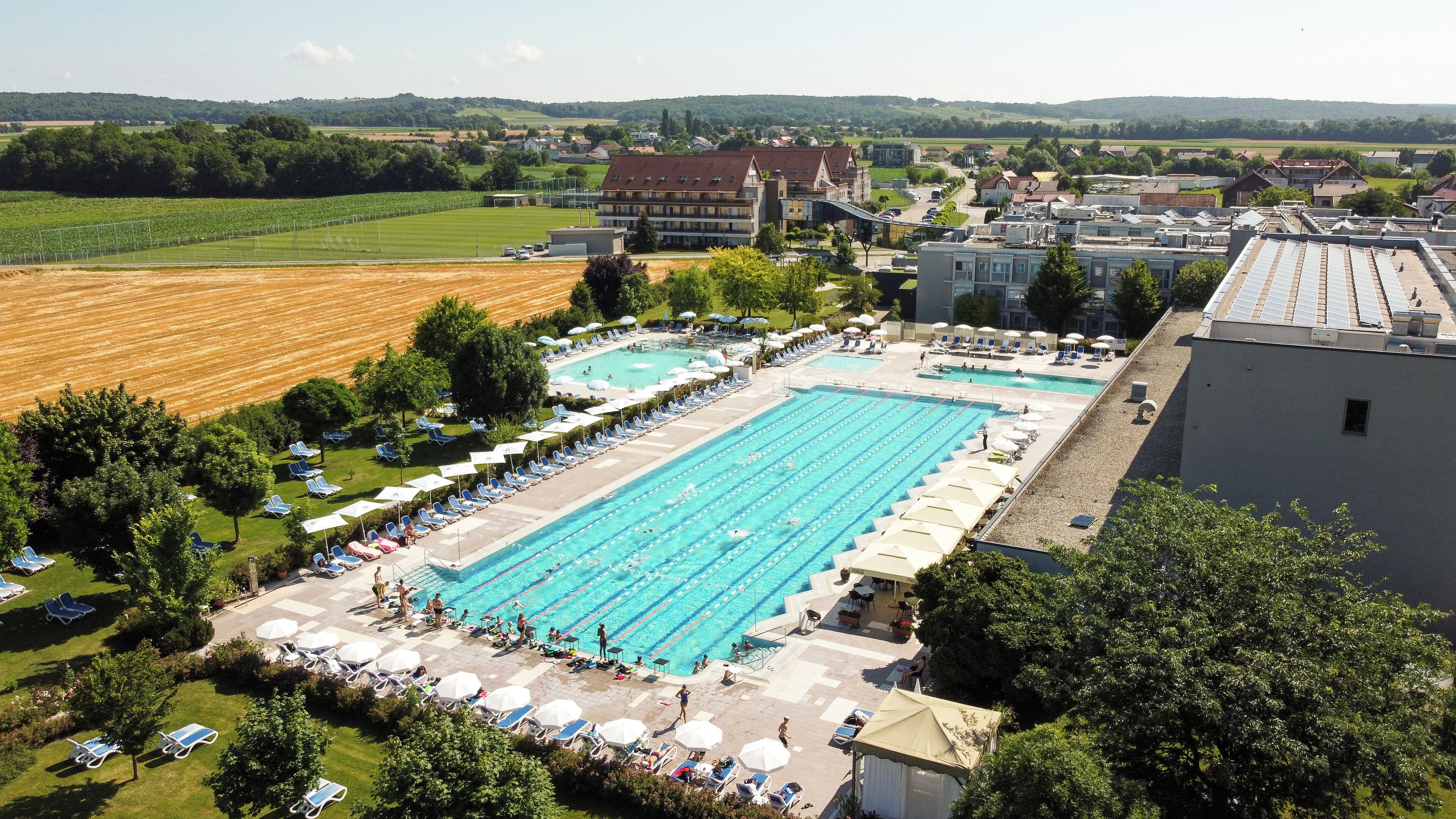 Uma grande piscina com várias pistas de natação, rodeada por espreguiçadeiras e guarda-sóis, com edifícios residenciais e campos abertos ao fundo.