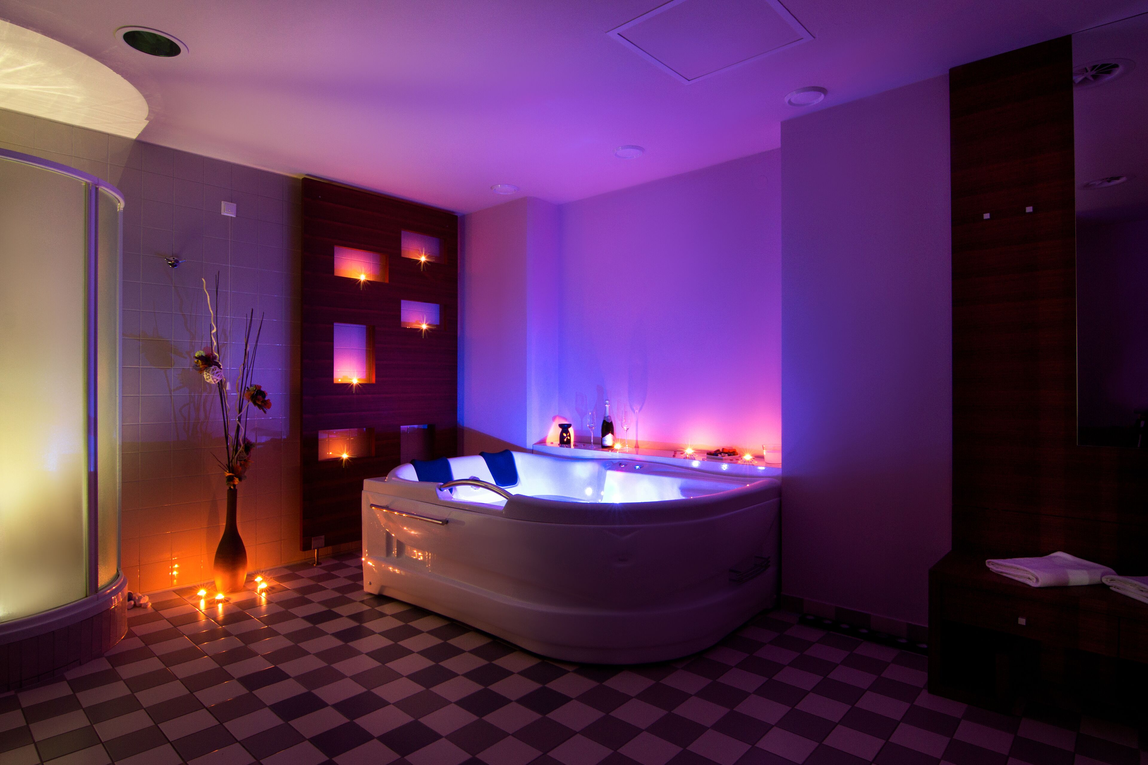 A modern bathroom with a checkered floor, a large bathtub with built-in lighting, a wooden wall panel, and a vase with flowers.