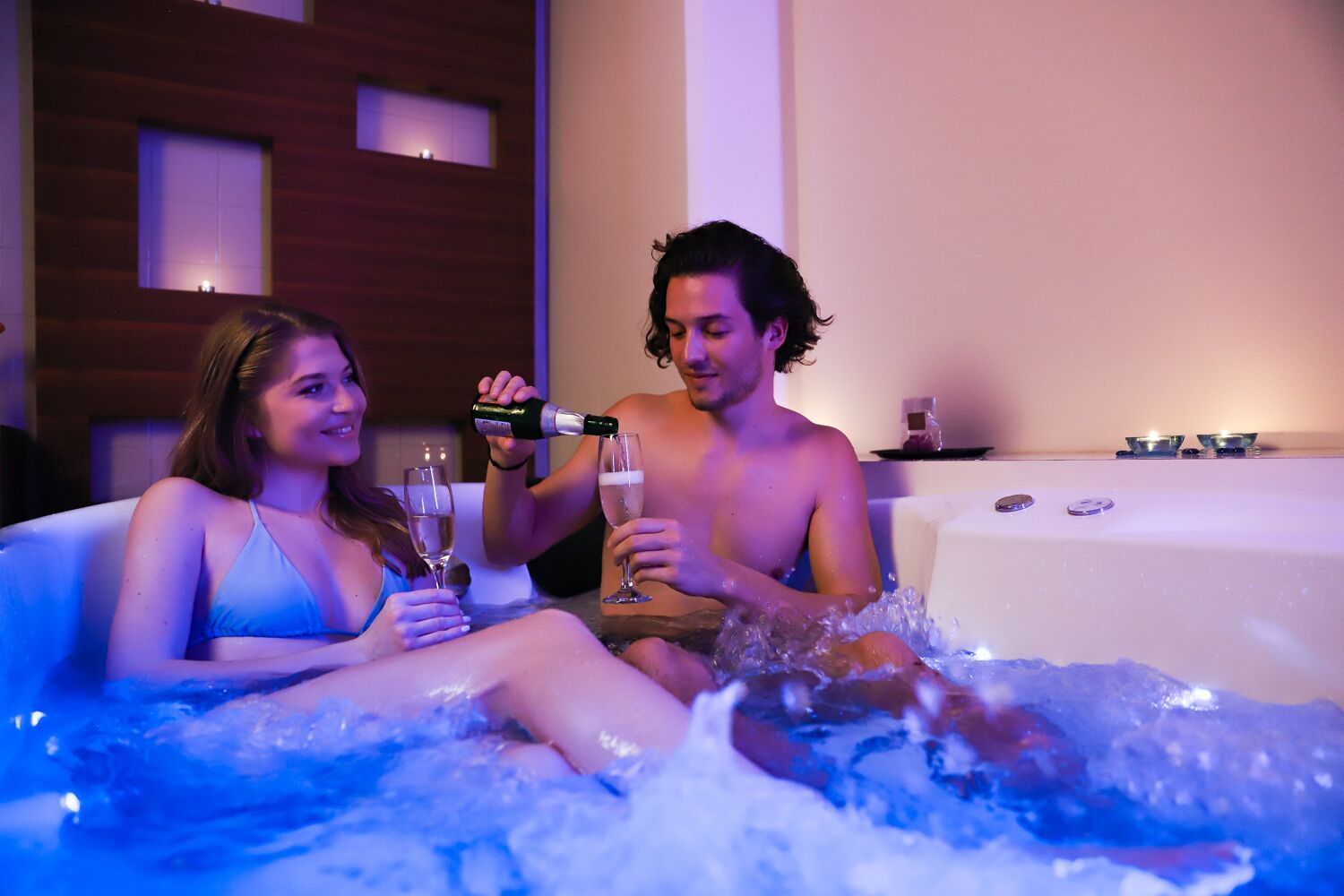 Dos personas en un jacuzzi con agua, sosteniendo copas de un líquido transparente, probablemente champán, en una habitación con paneles de madera y una ventana pequeña.