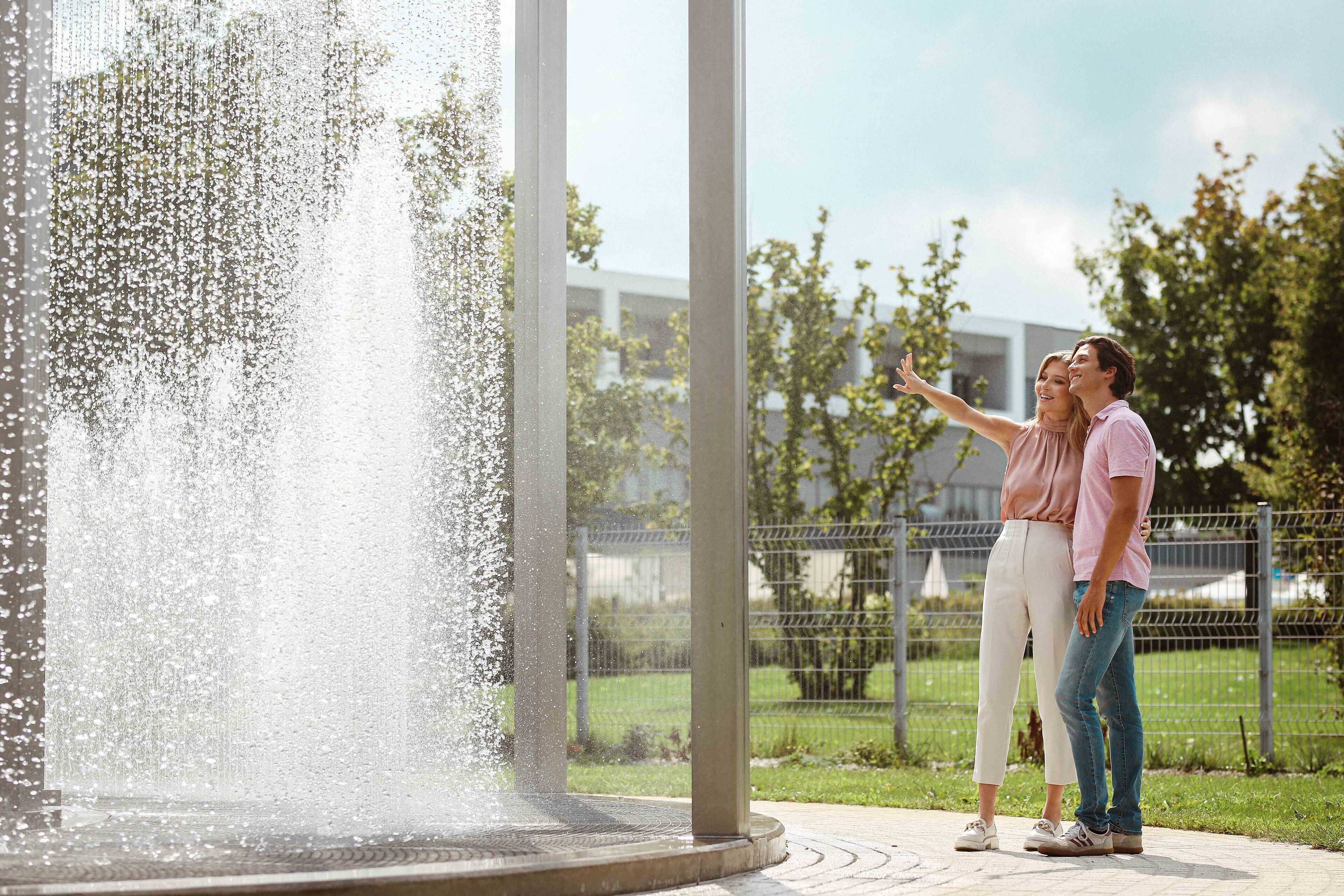 A couple standing by a fountain with a modern building in the background.