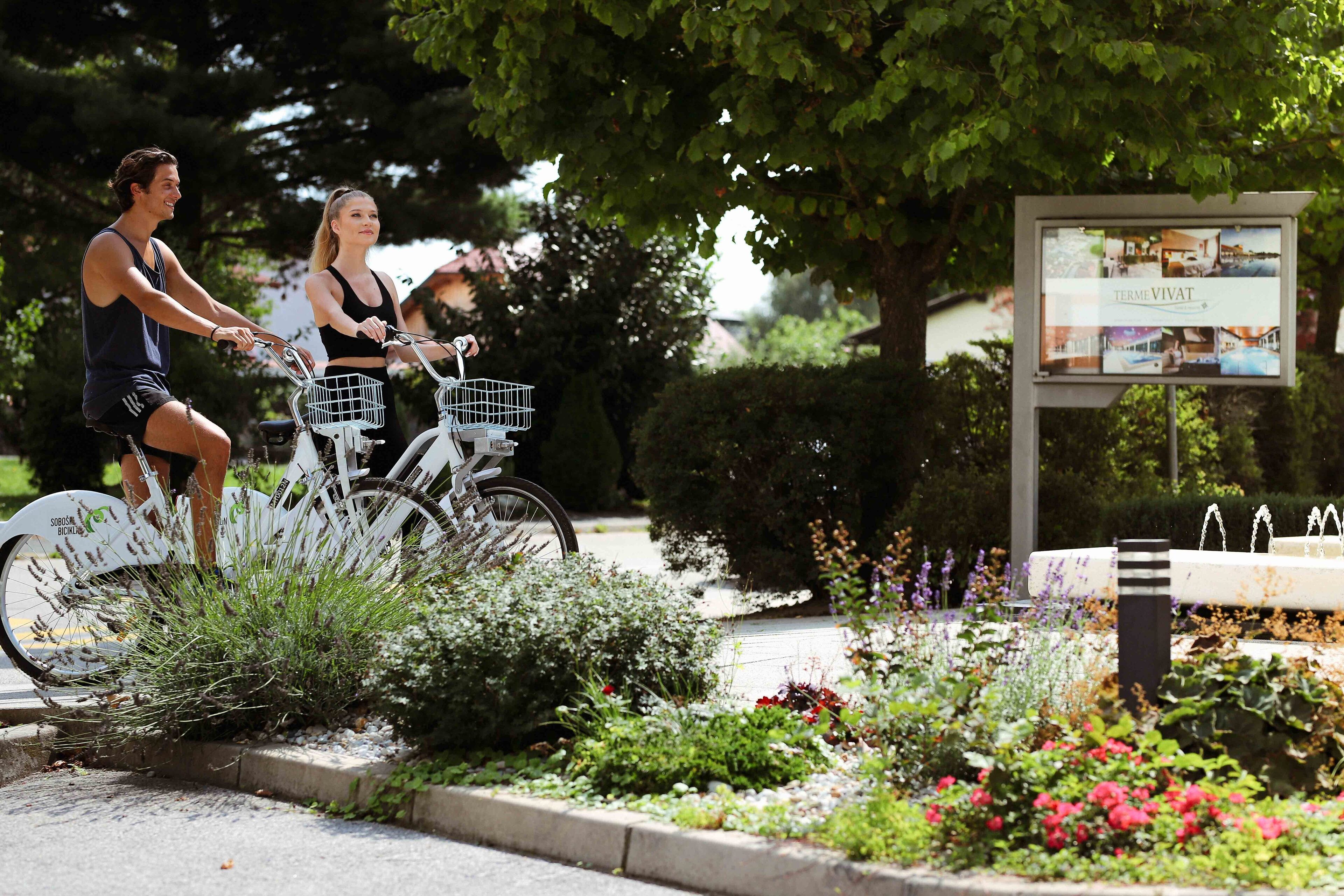 Dos personas en bicicleta, un jardín con flores y un cartel.