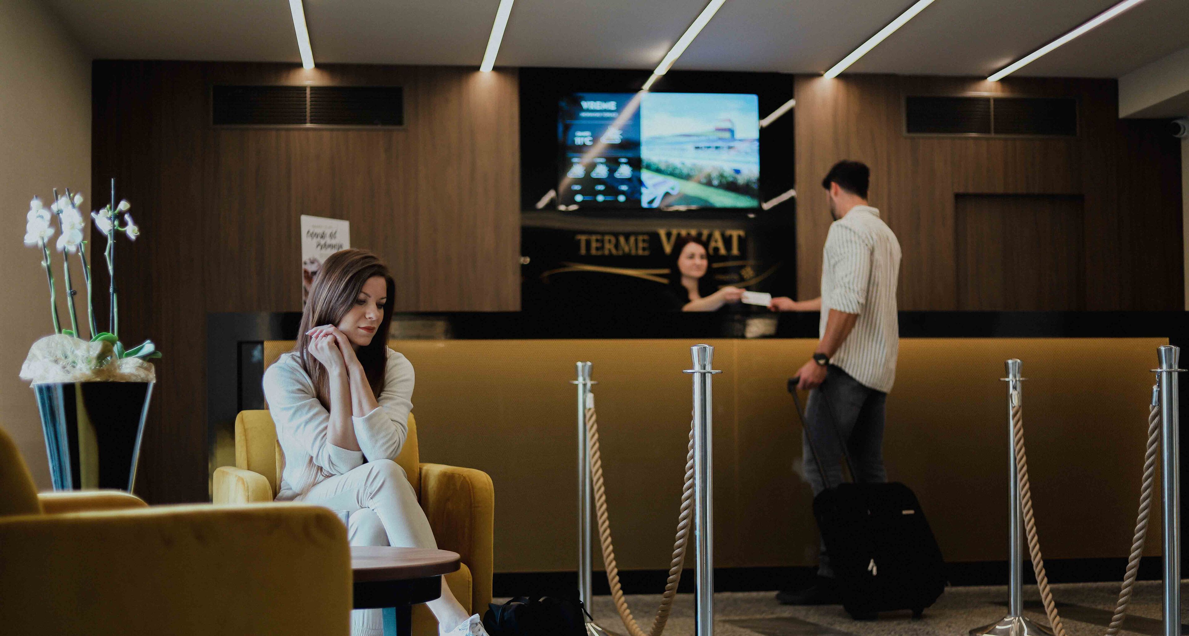 Zona de recepción de hotel con mostrador, área de espera con un sillón amarillo y una pantalla de televisión que muestra un paisaje urbano.