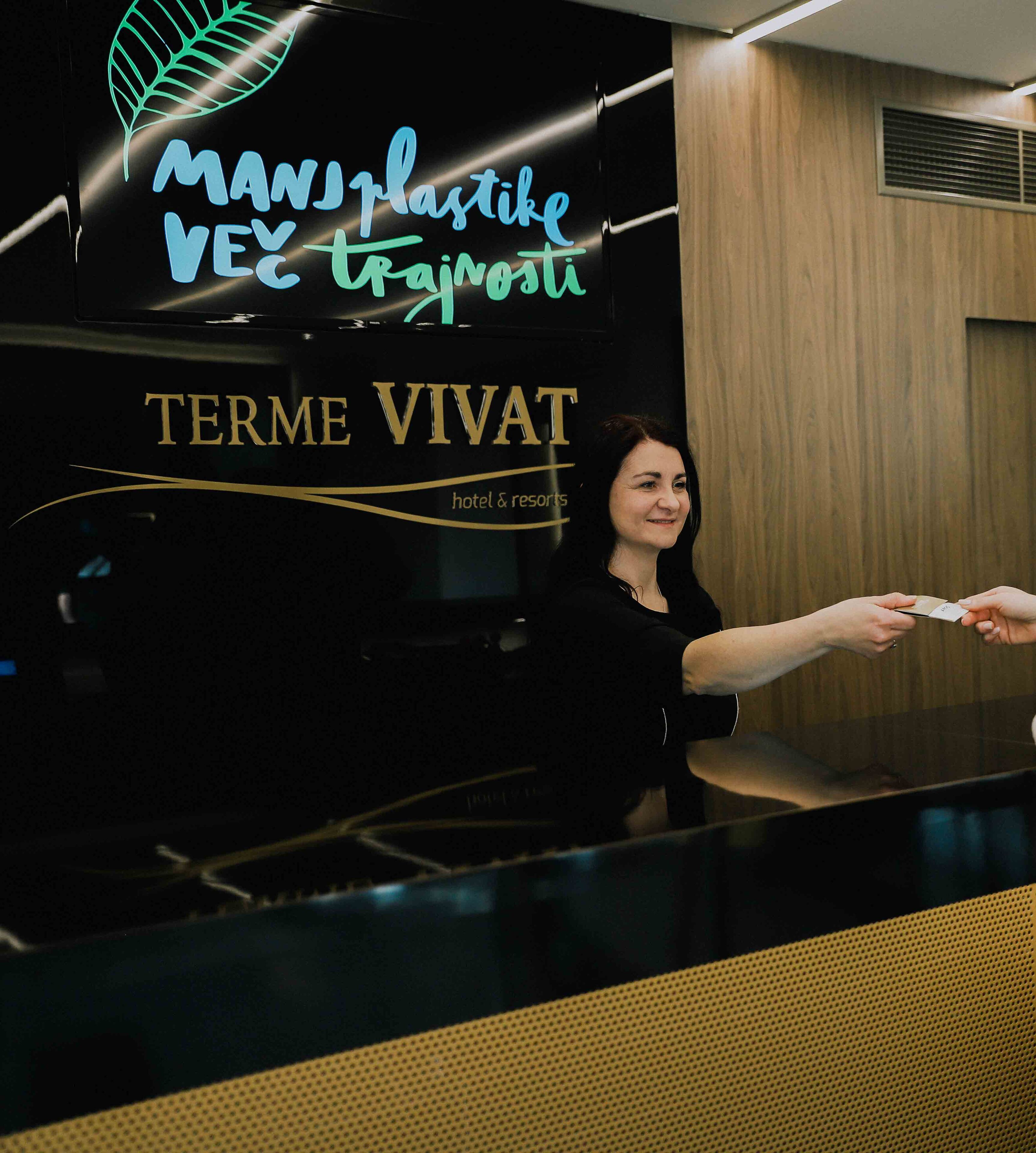 A woman at a reception desk with a sign above that reads "MANV Plastike VEC Trgovsti" and "TERME VIVAT hotel & resorts".
