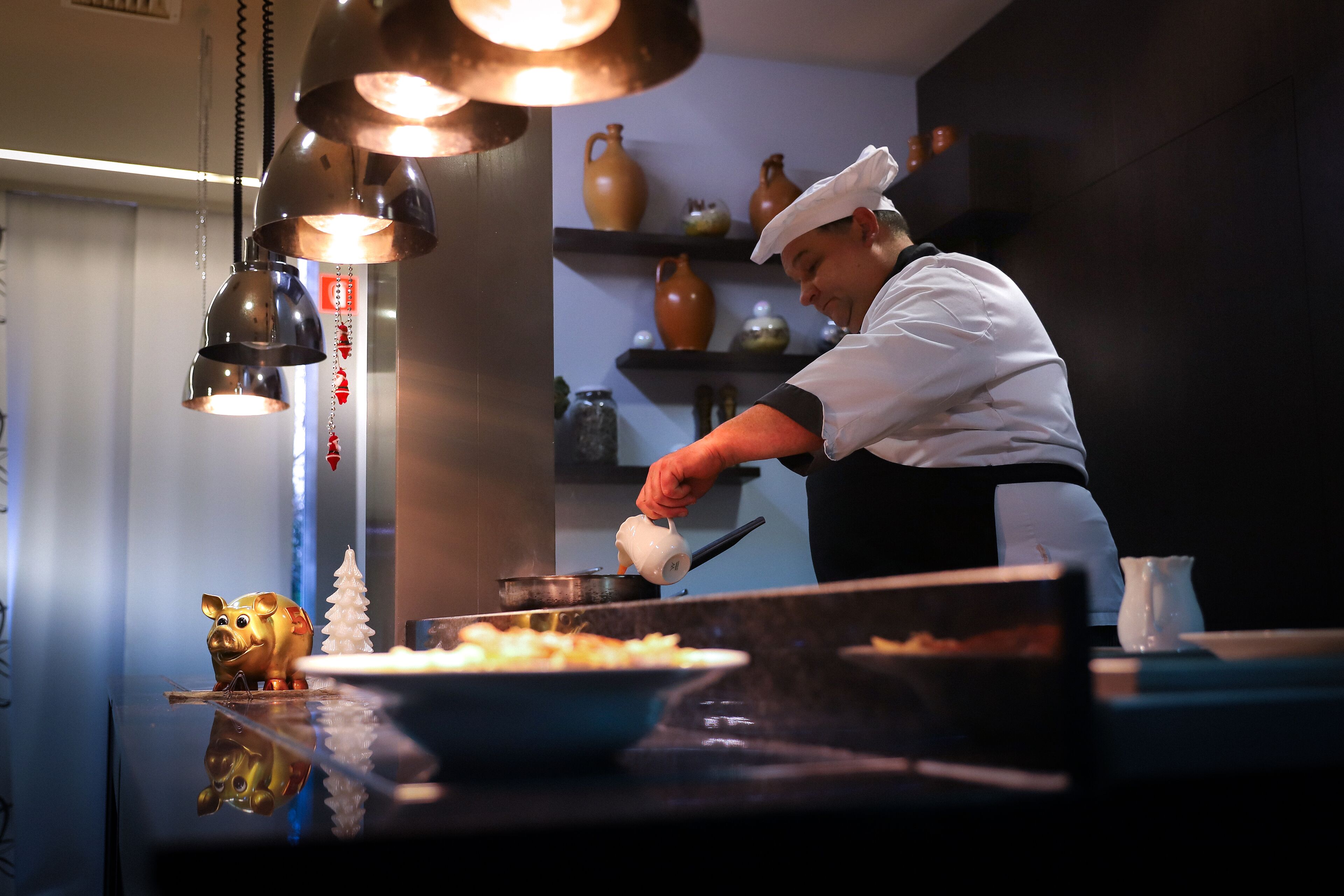 Un chef en una cocina profesional con luces colgantes, un cerdito alcancía y jarrones decorativos en estantes.