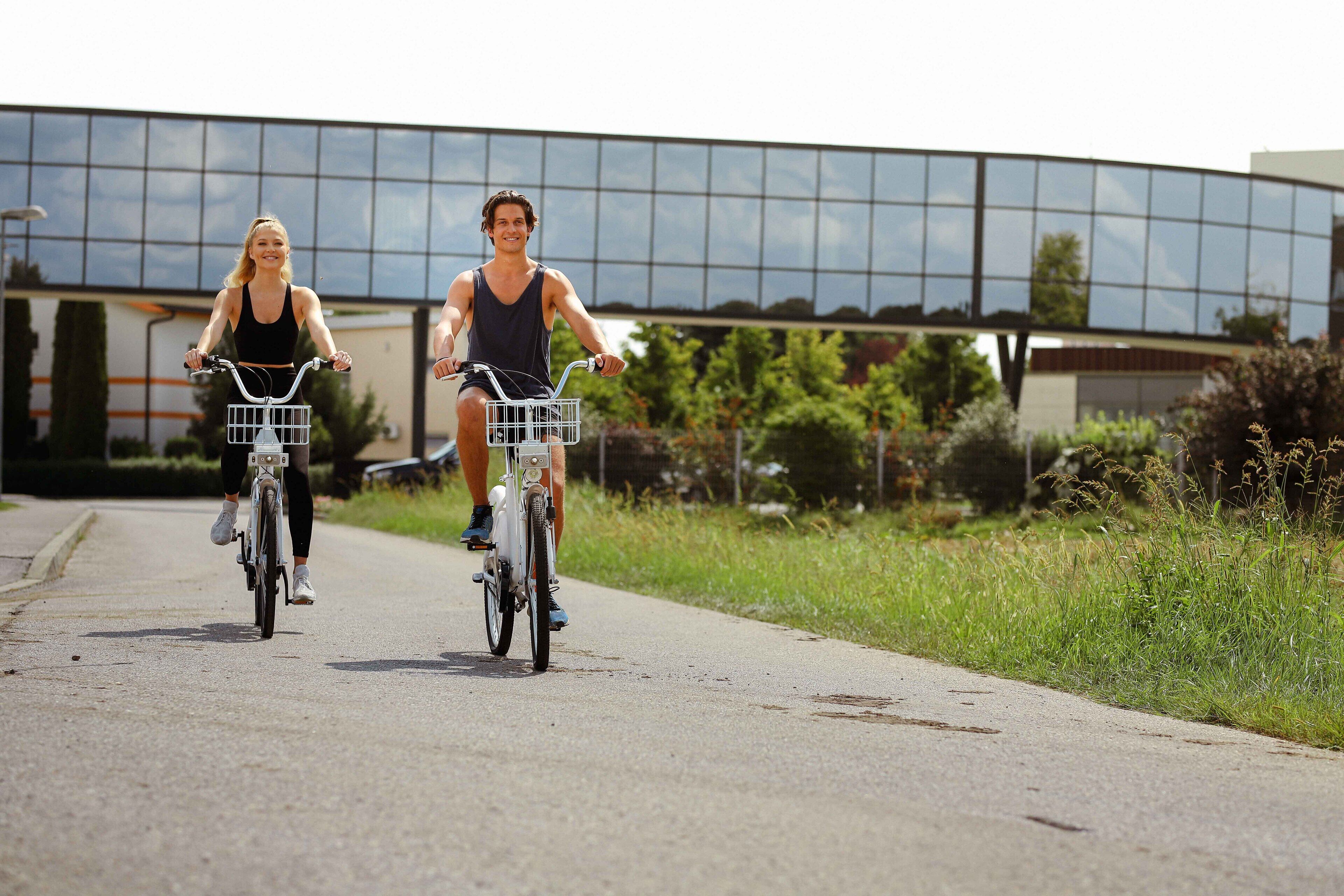 Two people riding bicycles on a paved path with a modern building in the background.