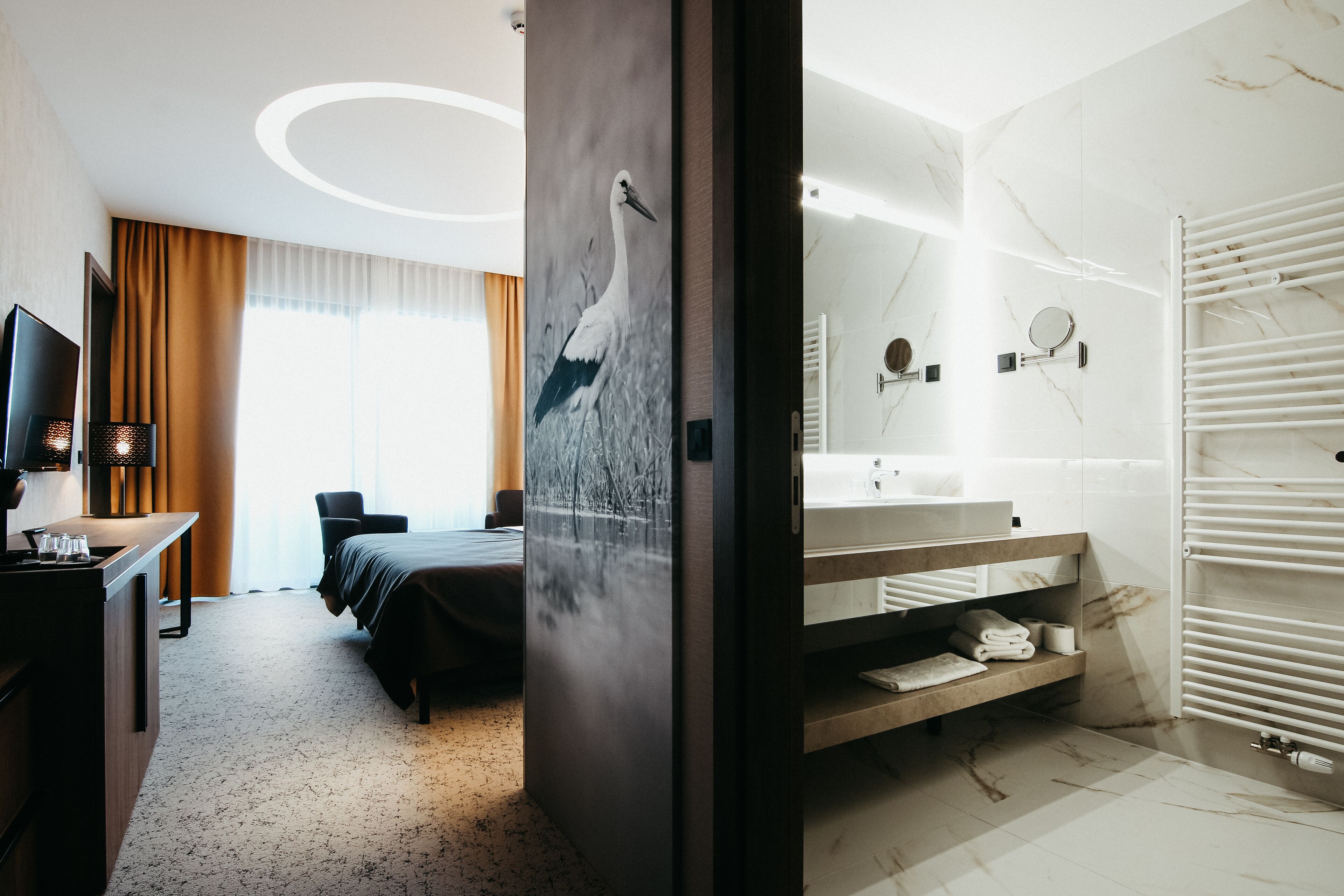 Habitación de hotel con cama, escritorio con computadora, ventana grande con cortinas, baño con lavamanos y espejo, y radiador fijado a la pared.