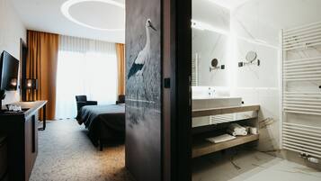 Habitación de hotel con cama, escritorio con computadora, ventana grande con cortinas, baño con lavamanos y espejo, y radiador fijado a la pared.