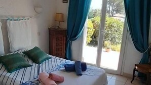 Makuuhuoneessa on sänky, kattotuuletin, ikkuna, jossa on verhot, ja ikkunasta avautuu näkymä vehreään maisemaan.