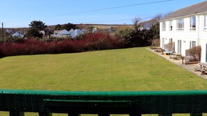 Un césped verde con una terraza de madera y mesas de picnic, rodeado de arbustos rojos y un edificio blanco con varias ventanas.