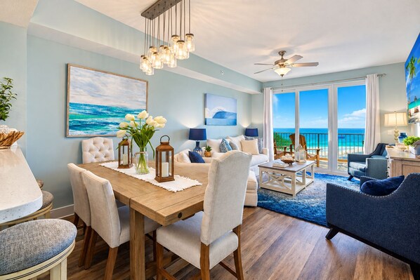 A spacious living area with a view of the ocean, a dining table with chairs, a ceiling fan, and a balcony with a view of the beach.