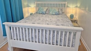 A bedroom with a white bed, patterned bedding, teal curtains, and two wall art pieces.