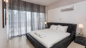 A modern hotel room with a large bed, a bedside table with a lamp, and a window with sheer curtains. -  Bay View Gardens Apts with Pool by TrulyCyprus (Limassol)