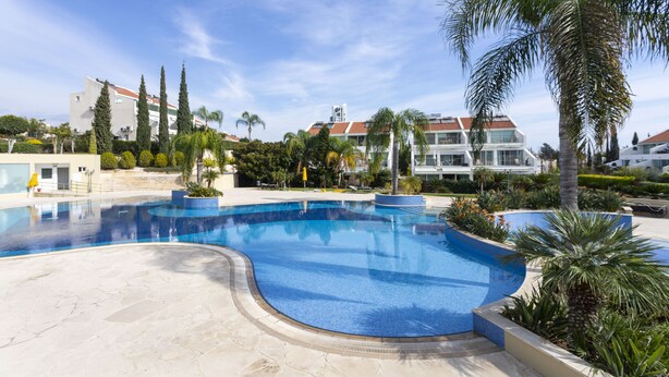 A swimming pool with a curved design, surrounded by palm trees and other greenery, with buildings in the background. -  Bay View Gardens Apts with Pool by TrulyCyprus (Limassol)