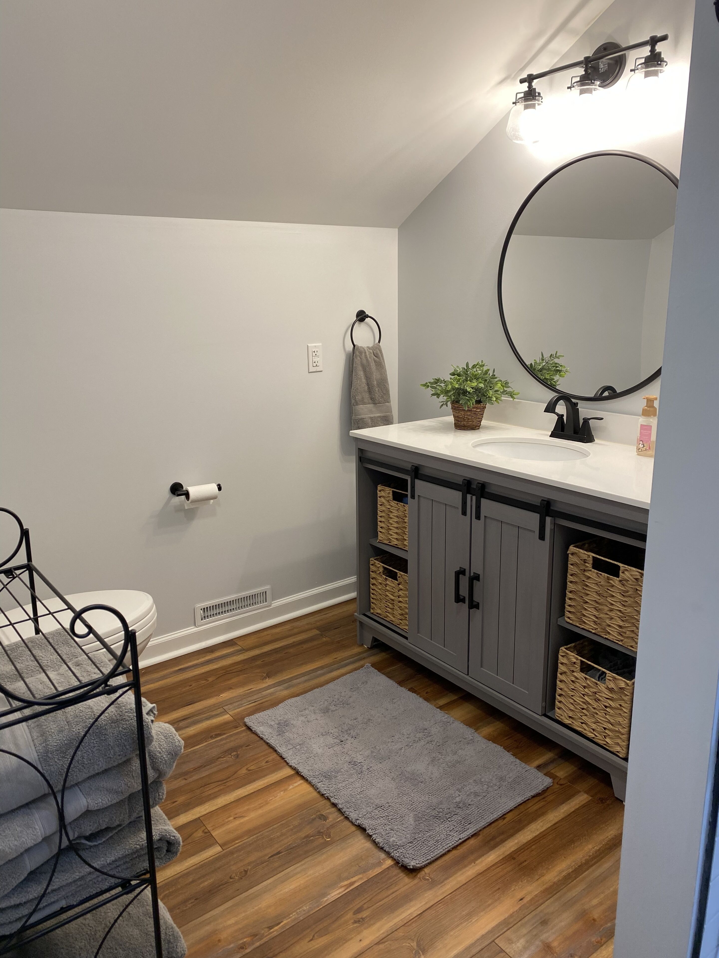 A bathroom with a toilet, a sink with a mirror, a towel rack, a basket, and a wooden floor.