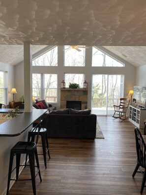A spacious living area with a high ceiling, large windows, a fireplace, and a kitchen area with bar stools.