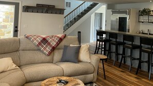 Una sala de estar con sofá modular, una mesa de centro, alfombra estampada, una escalera y una zona de cocina con taburetes de bar.